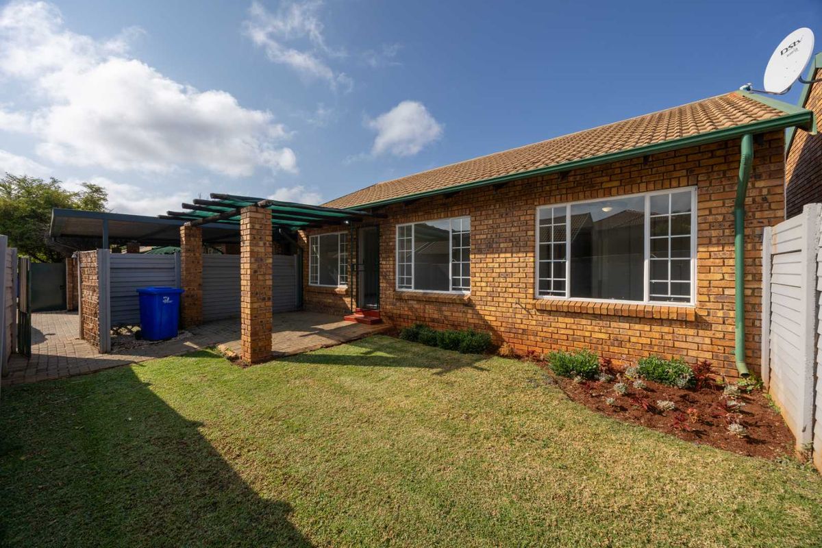 Rear view of the townhouse featuring a brick facade, tiled roof, paved patio area with a pergola, and a well-maintained garden lawn.