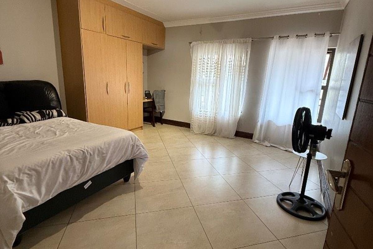 Spacious bedroom featuring neutral tiled flooring, built-in wooden cupboards, and large windows with curtain rails, offering a functional and well-lit living space.