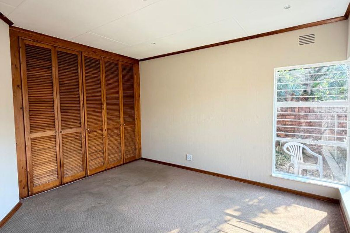 A spacious, empty bedroom featuring built-in wooden louvered cupboards, carpeted flooring, and natural light through a window with security bars.
