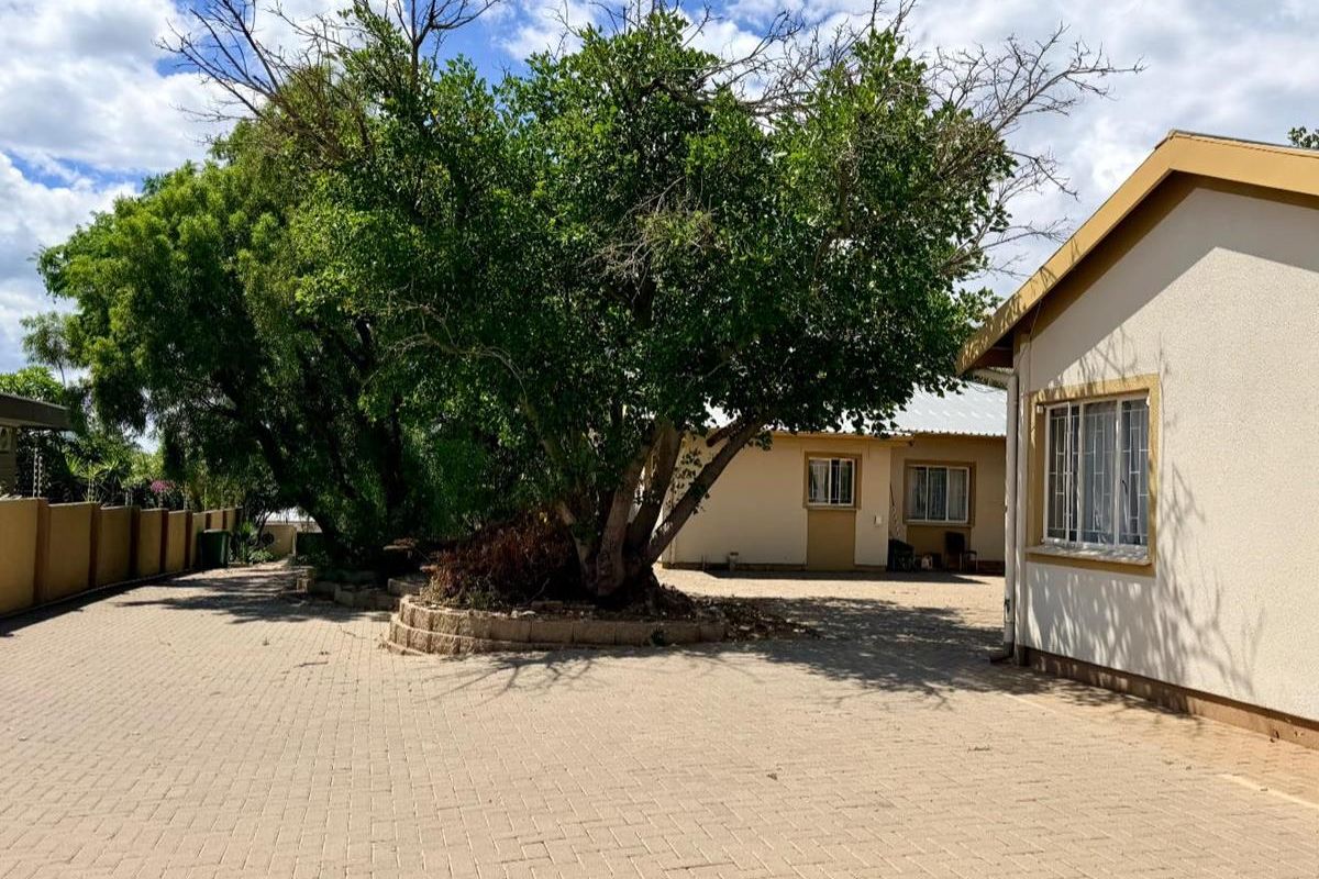 A paved driveway and courtyard area featuring a large central tree, perimeter boundary walls, and exterior access to the main house structure.