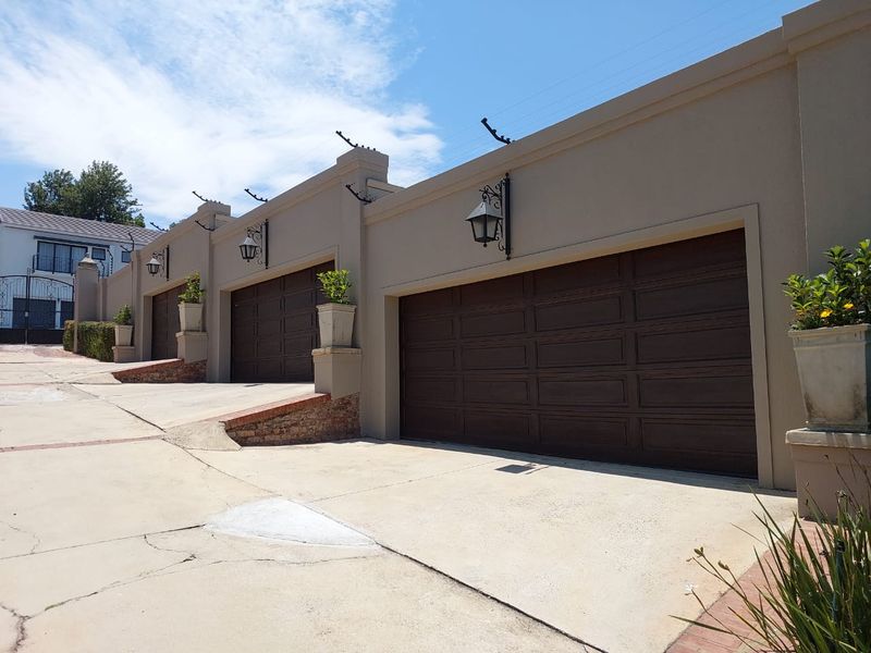 A triple garage exterior featuring dark wood-finish sectional doors, wall-mounted lanterns, and secure electric fencing along the perimeter wall.