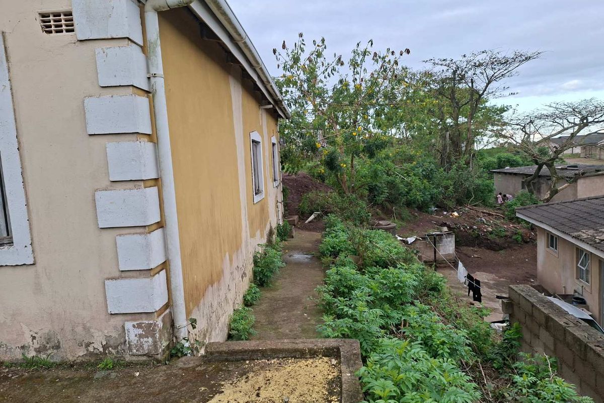 Rear exterior view of the property showing the side facade, concrete pathway, and surrounding garden area with visible signs of wear and maintenance needs.