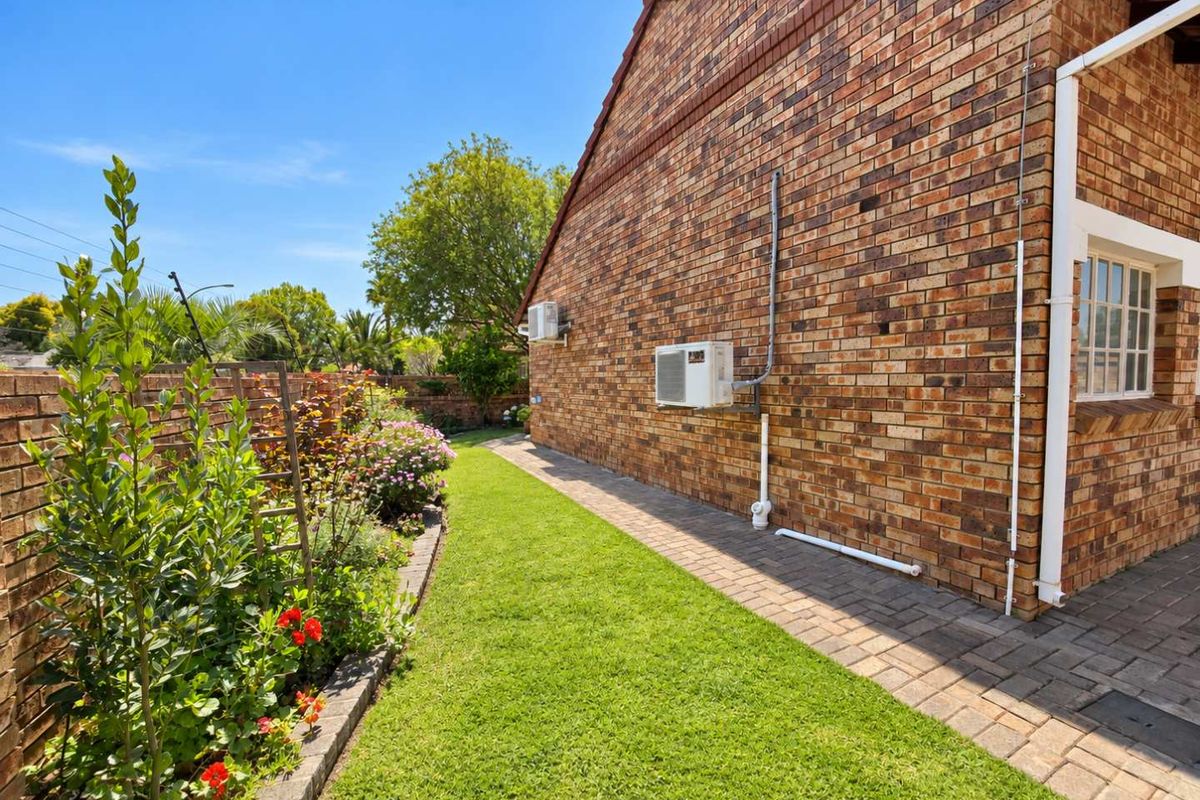 A well-maintained rear garden area featuring a paved walkway, lush lawn, and face brick exterior wall with an installed air conditioning unit.