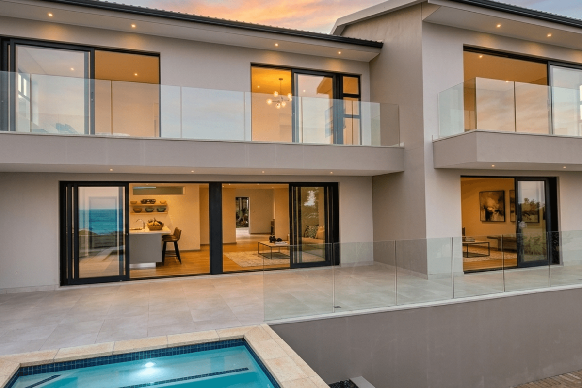 Modern rear elevation featuring a swimming pool, expansive tiled patio, glass balustrades, and large sliding doors opening into the living and kitchen areas.
