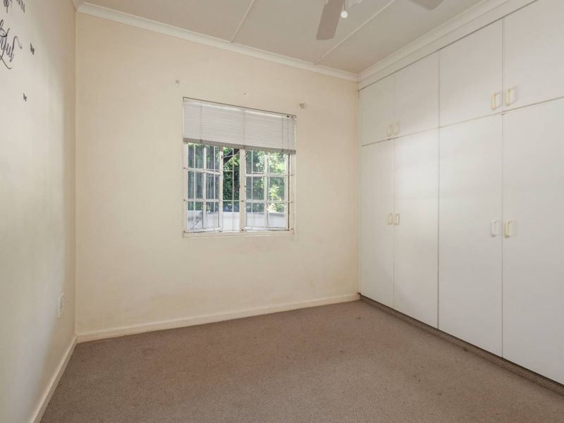 An empty bedroom featuring built-in cupboards, a ceiling fan, and security bars on the window, with carpeted flooring throughout.
