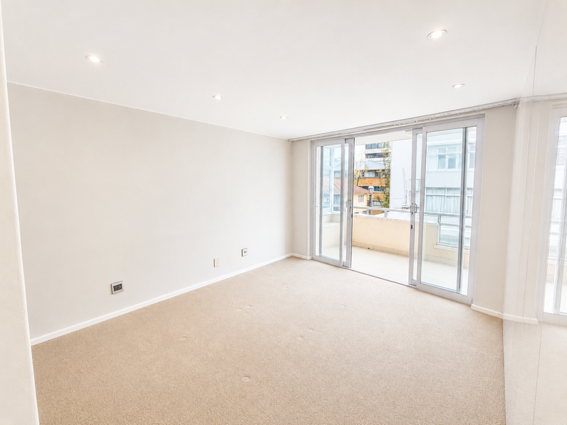 A bright, empty living room featuring neutral carpeting, recessed lighting, and large sliding glass doors that open onto a private balcony.