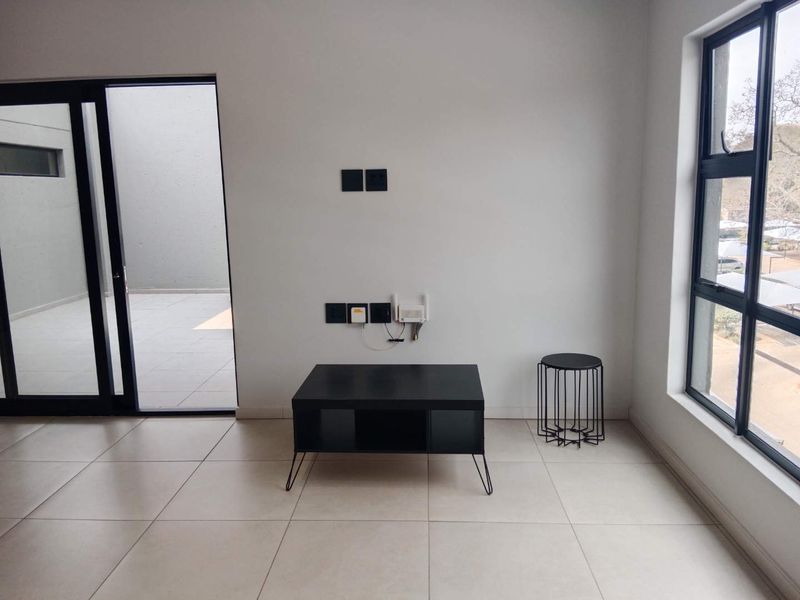 A bright, modern living area featuring neutral tiled flooring, large aluminium-framed windows, and direct access to an outdoor patio space.