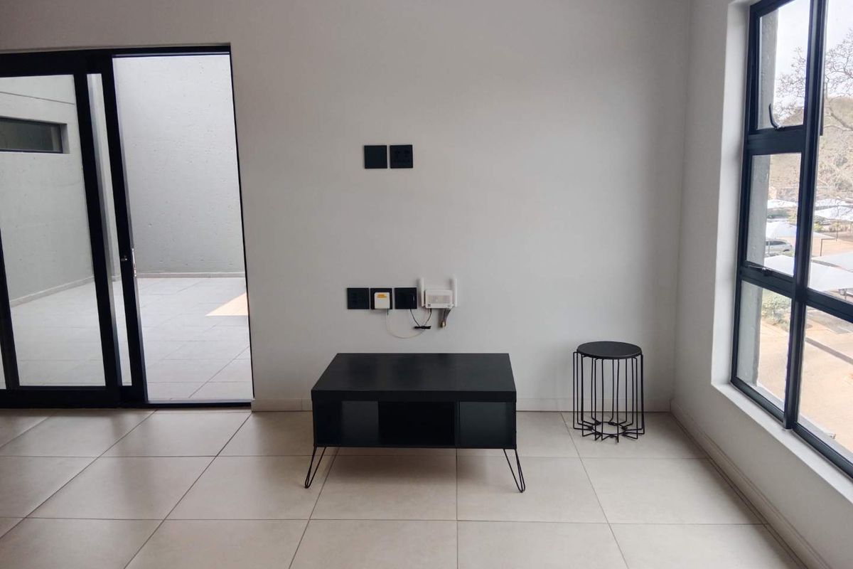A bright, modern living area featuring neutral tiled flooring, large aluminium-framed windows, and direct access to an outdoor patio space.