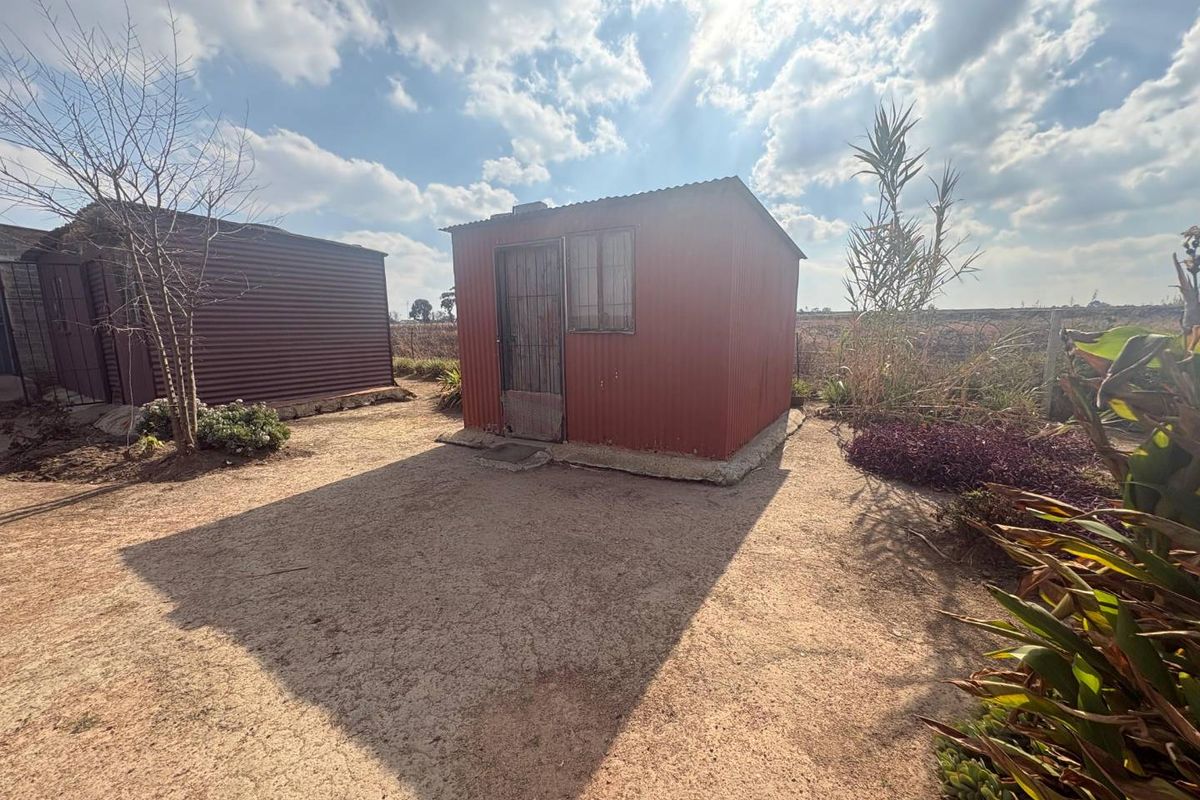 A small, detached corrugated metal structure situated on a vacant residential plot, featuring a simple door and window configuration.
