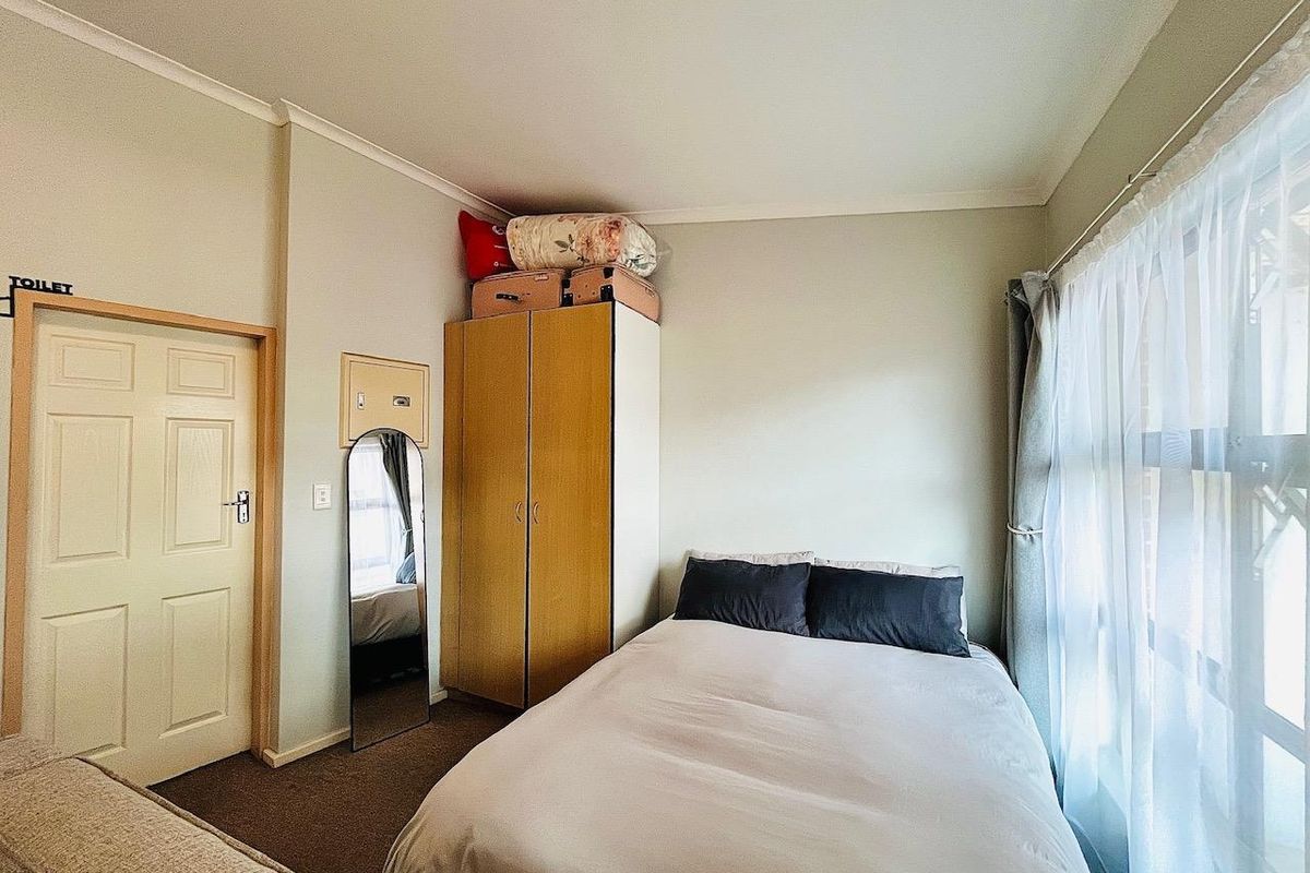 A functional bedroom featuring neutral wall finishes, a built-in cupboard, and natural light provided by a large window with curtain rails.