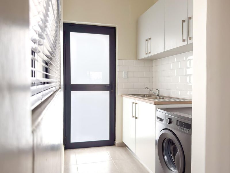 A clean and functional laundry area featuring built-in white cabinetry, a stainless steel sink, subway tile backsplash, and direct access to the exterior.