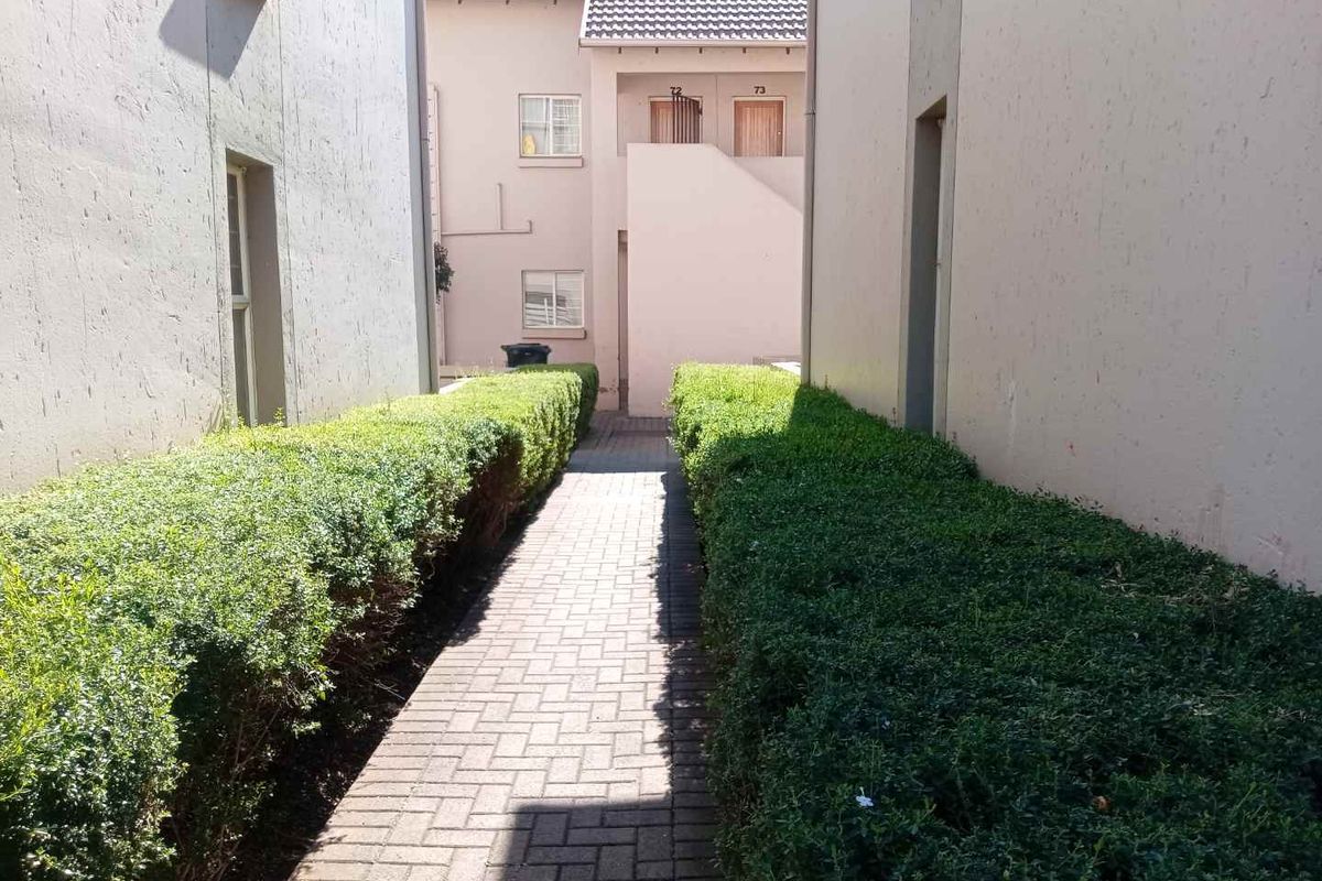 A paved walkway leading between two apartment building sections, flanked by manicured hedges.