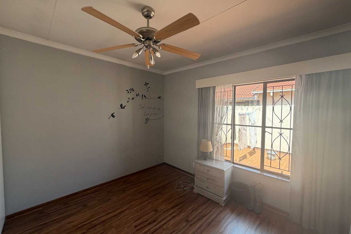 A bedroom featuring wood-look laminate flooring, a ceiling fan, and a window with security bars overlooking a paved outdoor area.