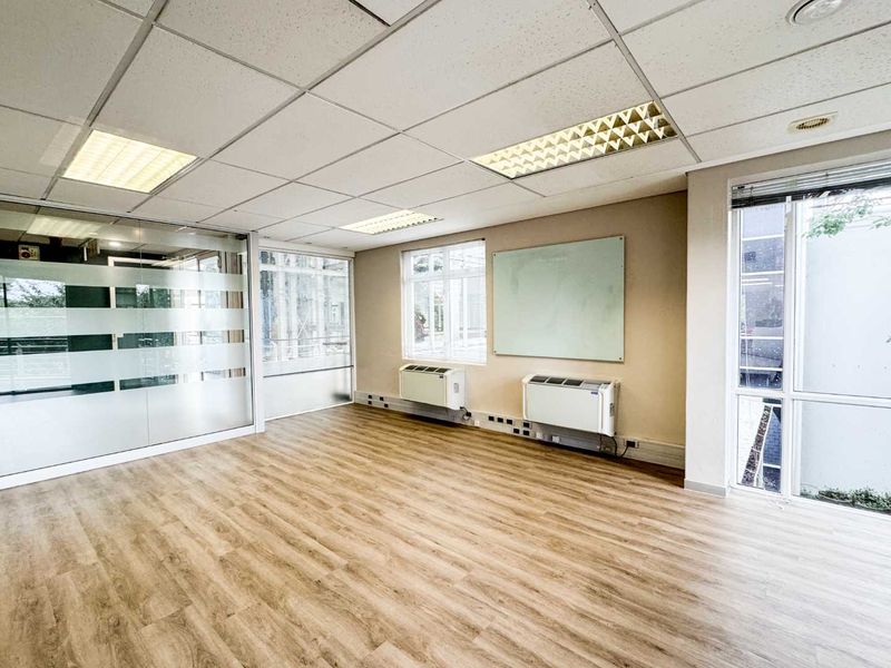 A bright, empty office space featuring laminate flooring, a glass partition, wall-mounted HVAC units, and a suspended ceiling with recessed lighting.