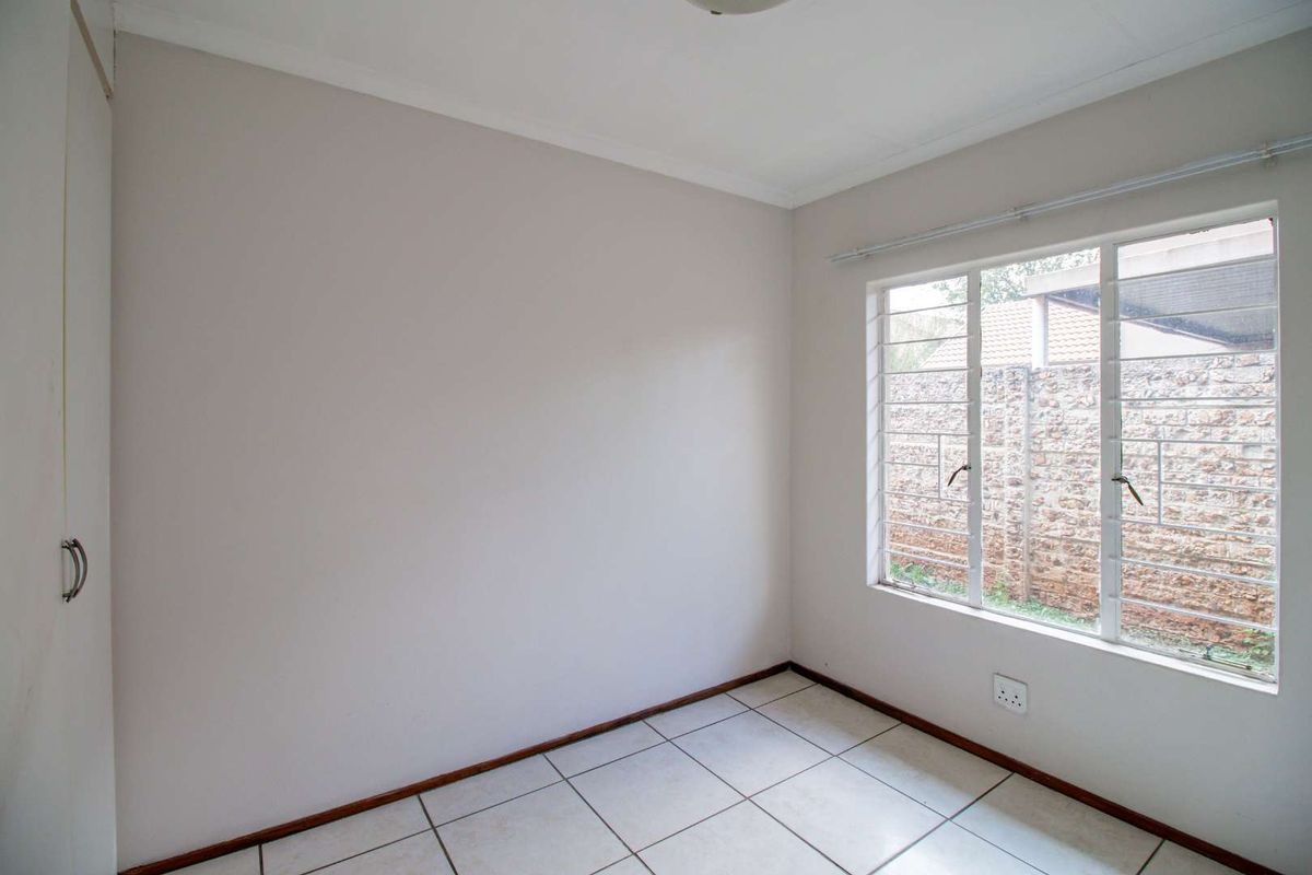 A bright, empty bedroom featuring neutral walls, tiled flooring, and a large window with security bars.