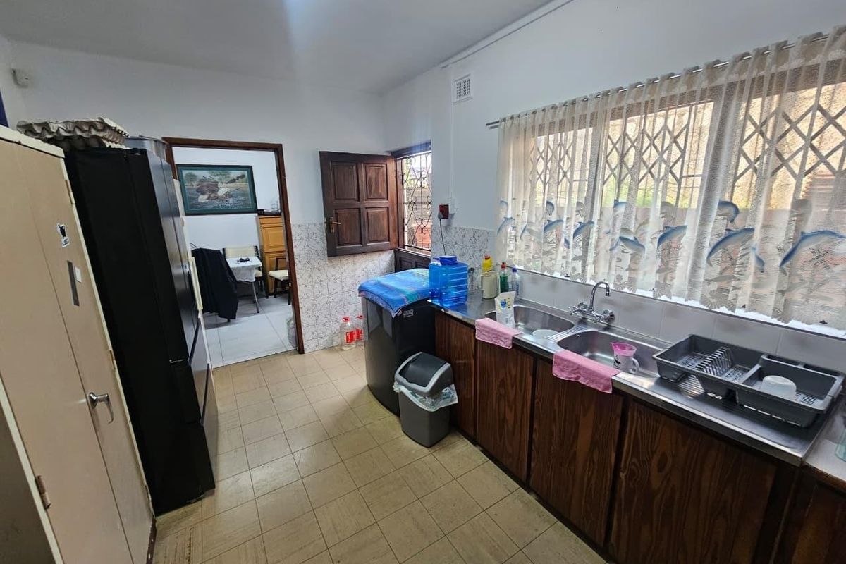 A functional kitchen featuring wooden cabinetry, a double bowl stainless steel sink, tiled splashbacks, and tiled flooring with access to an adjacent room.