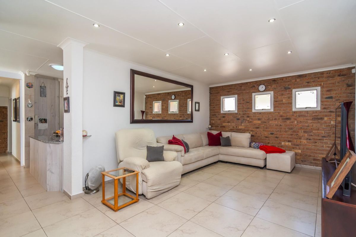 Spacious living room featuring an exposed brick feature wall, neutral tiled flooring, and recessed downlighting, with an open-plan flow to the adjacent kitchen area.