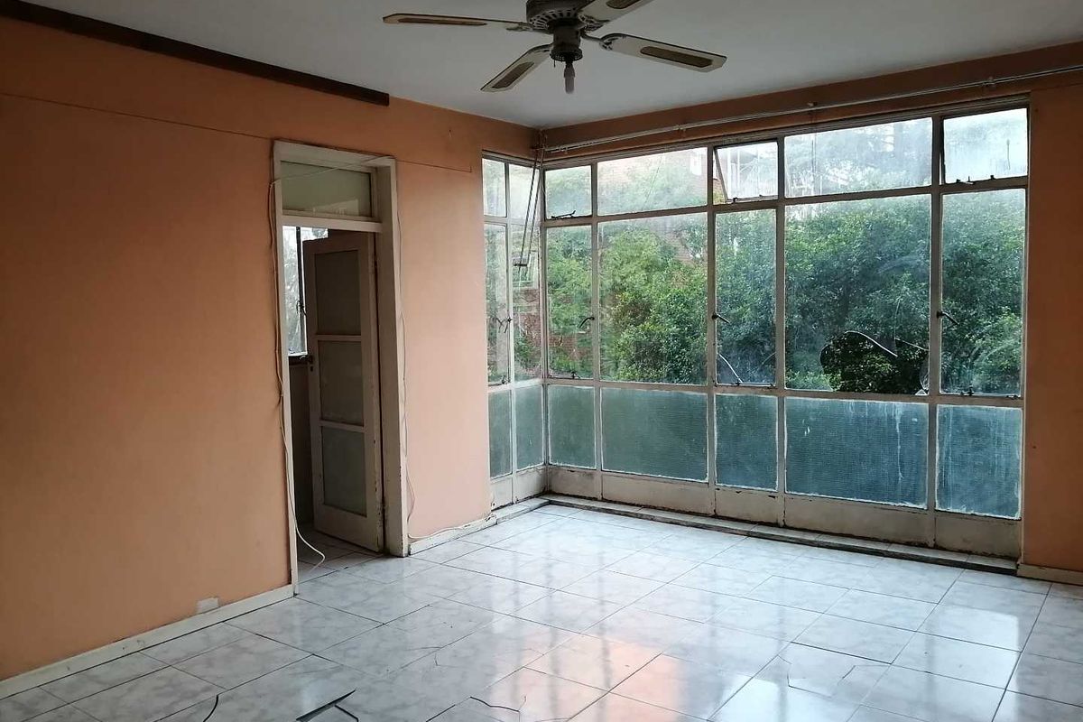 An empty living area featuring large floor-to-ceiling windows, tiled flooring, and a ceiling fan, offering a view of the surrounding greenery.