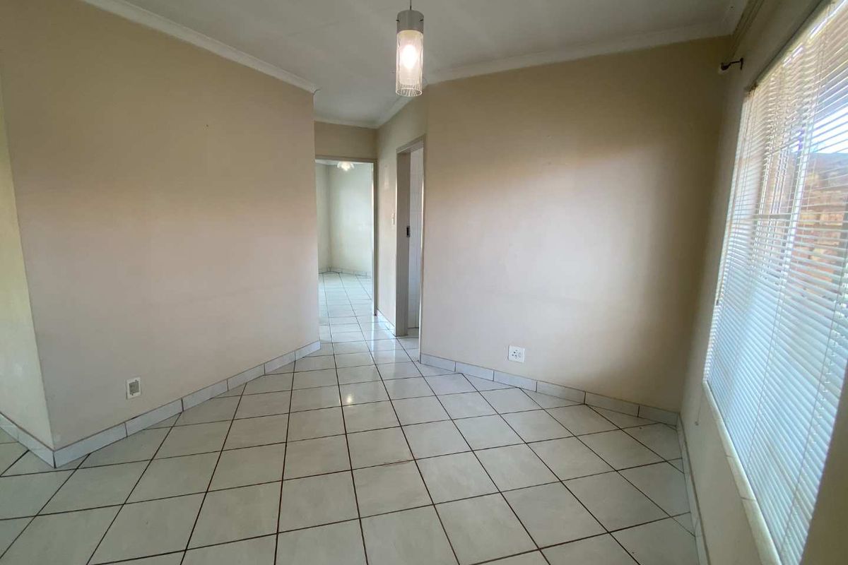 A bright, empty living area featuring neutral tiled flooring, a pendant light fixture, and large windows with fitted blinds, leading into a hallway.