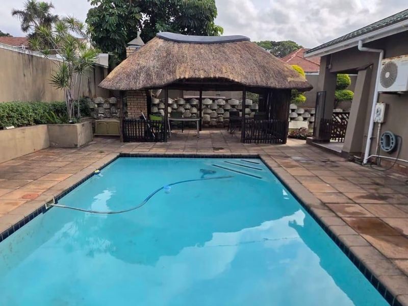 Outdoor pool area featuring a paved patio, a traditional thatched lapa for entertainment, and a perimeter retaining wall.
