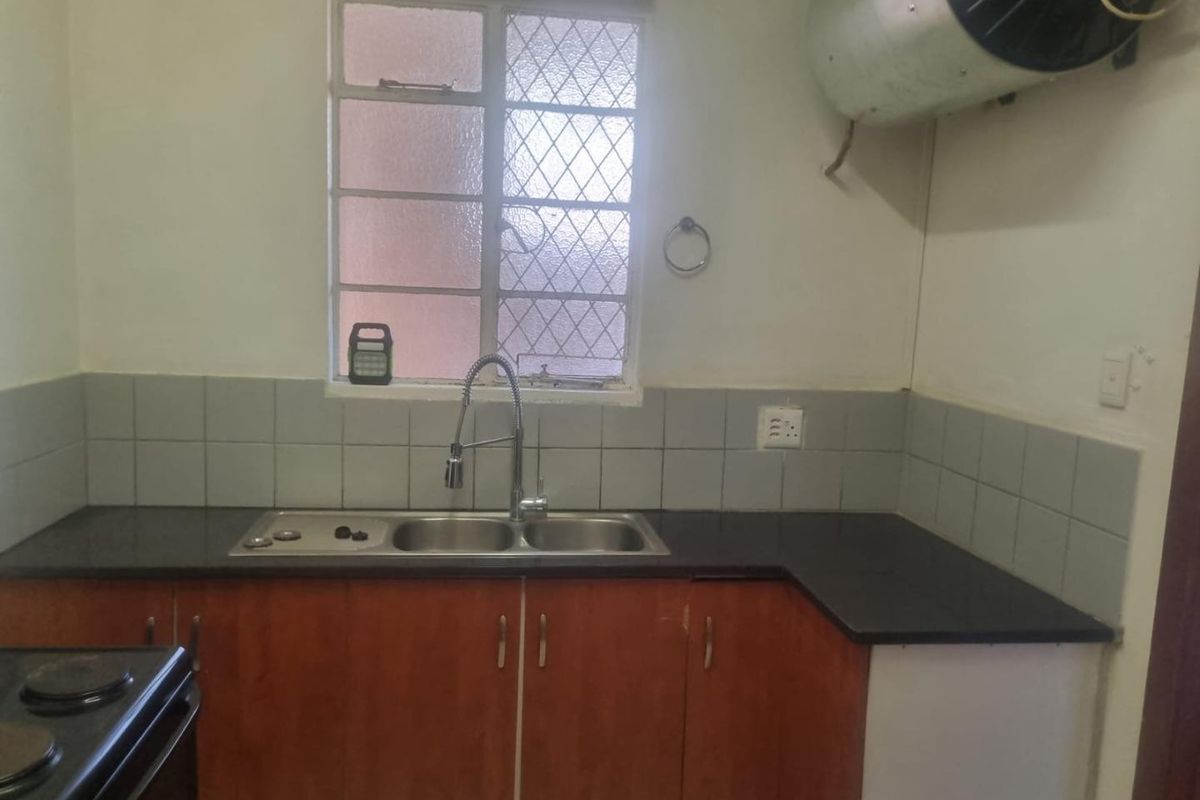A functional kitchen featuring dark stone countertops, double bowl sink, tiled splashback, and wooden cabinetry, with an exposed wall-mounted electric geyser.