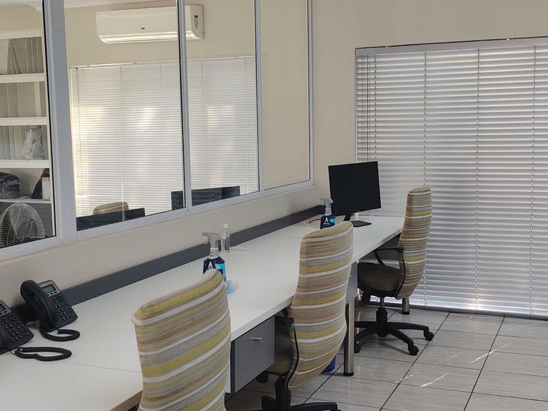 A functional office space featuring a long built-in desk, tiled flooring, split-system air conditioning, and large windows with venetian blinds.
