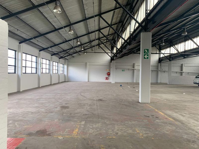 Spacious industrial warehouse unit featuring high exposed steel trusses, concrete flooring, ample natural light from large windows, and essential fire safety equipment.