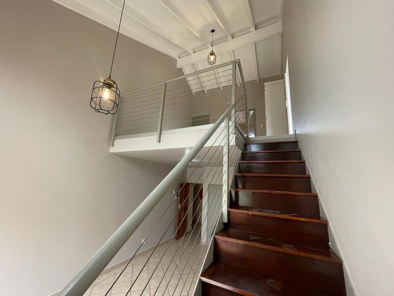 A bright hallway featuring a timber staircase with a modern metal balustrade, leading up to a mezzanine level with exposed beam ceilings.