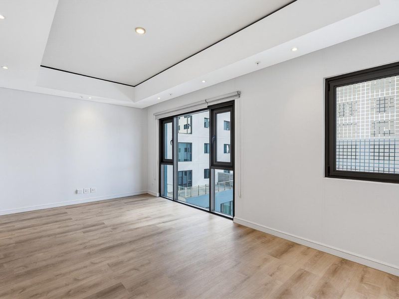 A bright, modern, and empty bedroom featuring light wood-look laminate flooring, a contemporary drop ceiling with recessed lighting, and large aluminium-framed windows.