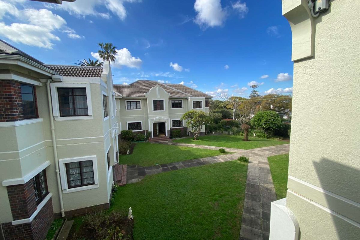A view of the apartment complex exterior featuring a well-maintained communal garden, paved pathways, and traditional brick and plaster building architecture.