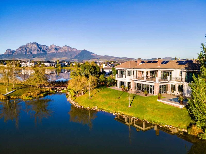 Aerial view of a luxury waterfront residence featuring a swimming pool, expansive patio, balcony, and solar panels set against a scenic mountain backdrop.