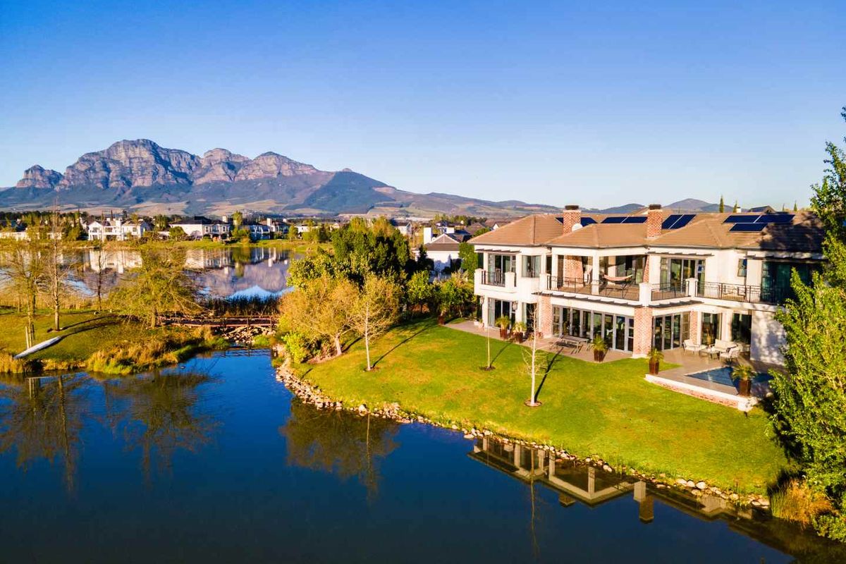 Aerial view of a luxury waterfront residence featuring a swimming pool, expansive patio, balcony, and solar panels set against a scenic mountain backdrop.