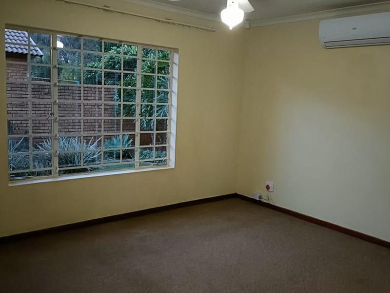 An empty bedroom featuring carpeted flooring, a ceiling fan, and a wall-mounted air conditioning unit with large windows and security bars.