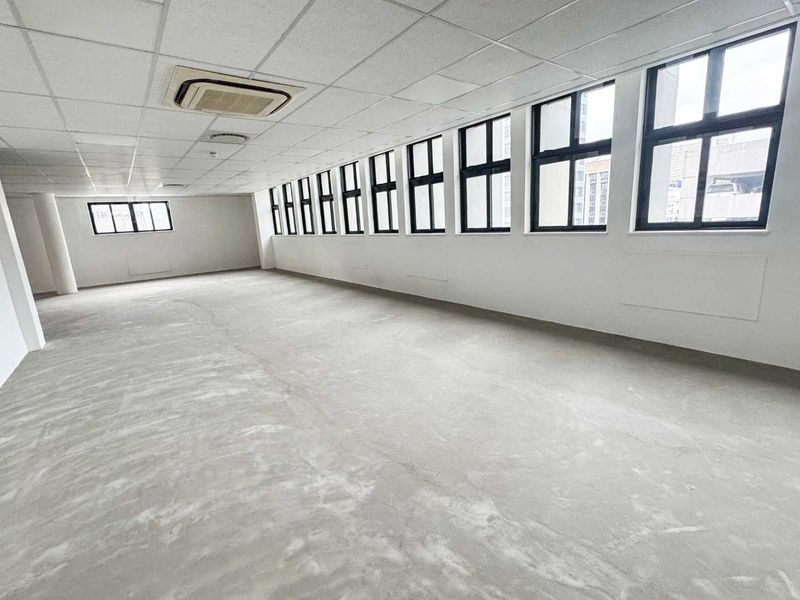 A spacious, empty open-plan office area featuring large windows, a suspended ceiling with integrated air conditioning, and unfinished concrete flooring.