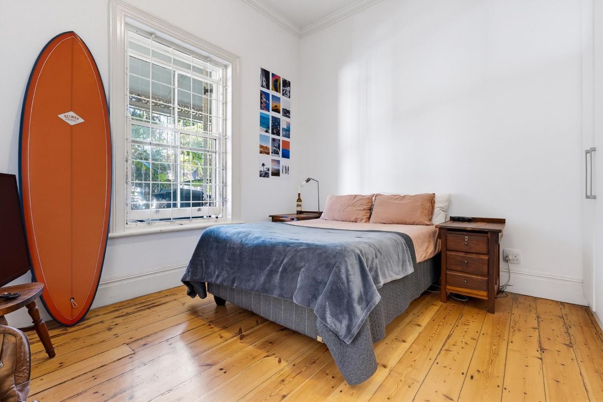 A bright bedroom featuring polished timber flooring, a large sash window with security bars, and classic cornice detailing.