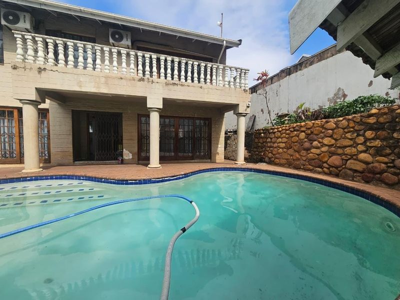 Rear exterior view featuring a swimming pool, paved patio area, and a two-story townhouse facade with a balcony and stone retaining wall.