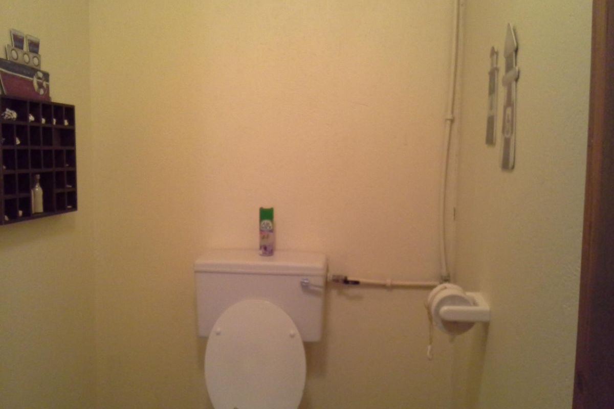 A small bathroom featuring a standard toilet with visible surface-mounted plumbing and a wall-mounted toilet roll holder.