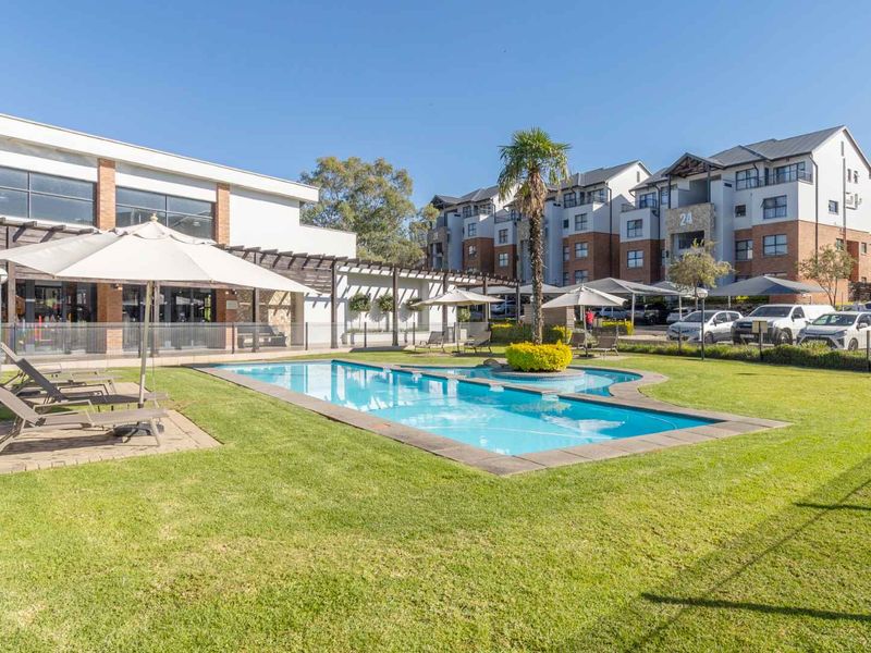 A well-maintained communal swimming pool area featuring a landscaped garden, paved deck, and pergola, situated within an apartment complex.