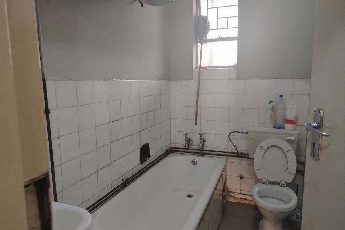 A dated bathroom featuring a standard bathtub, toilet, and basin with exposed plumbing and wall tiling in need of renovation.