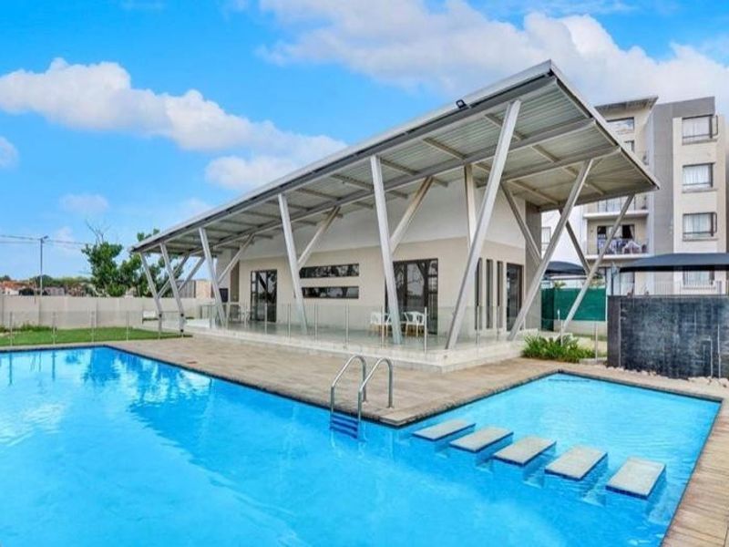 A modern communal pool area featuring a large swimming pool with stepped entry, a covered patio structure, and a contemporary water feature.