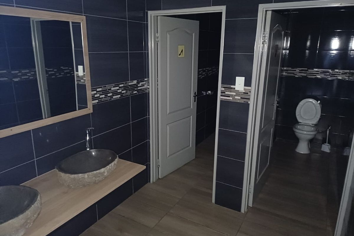 A modern bathroom featuring dual stone vessel sinks on a wooden vanity, dark tiled walls with decorative accents, and separate toilet cubicles.