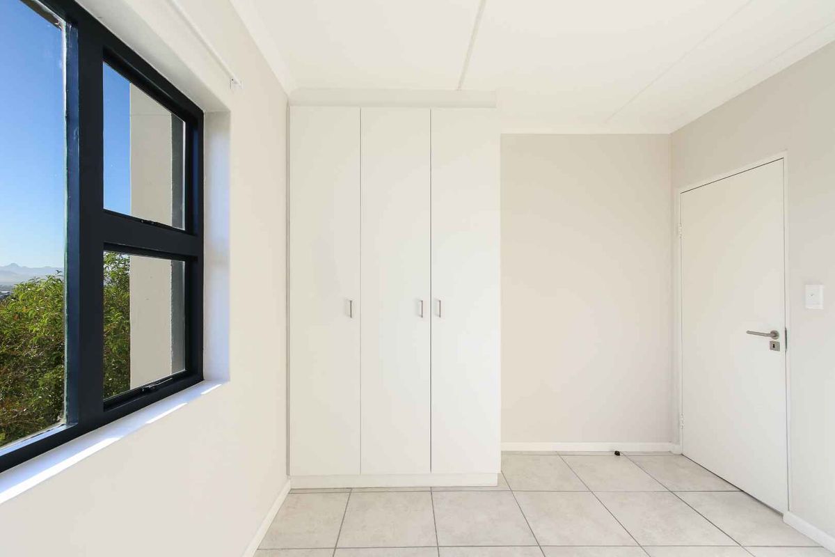 A bright, empty bedroom featuring neutral tiled flooring, built-in white cupboards, and a large window with views.