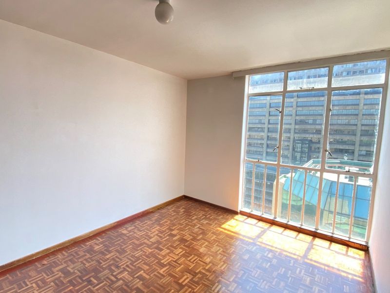 A bright, empty bedroom featuring classic parquet flooring and a large window with urban views, offering a clean canvas for personalization.