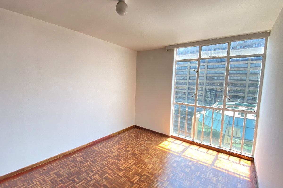 A bright, empty bedroom featuring classic parquet flooring and a large window with urban views, offering a clean canvas for personalization.