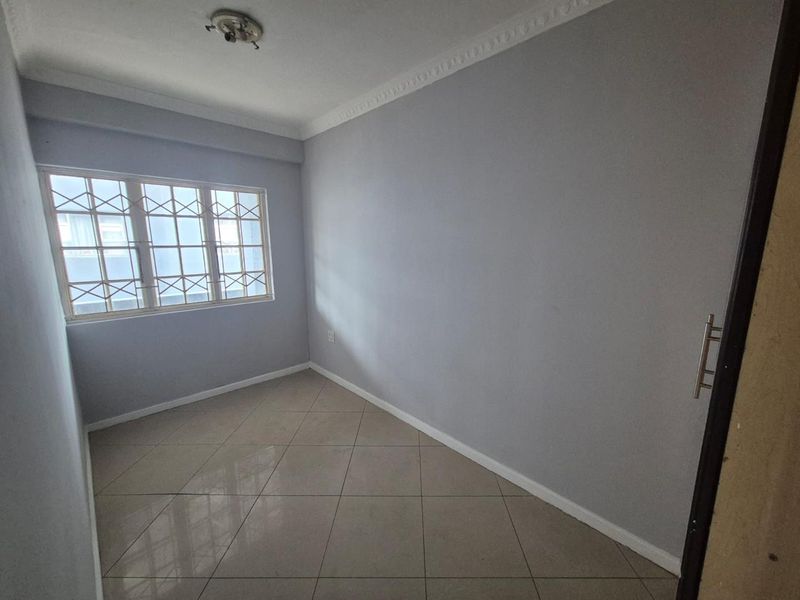 A clean, empty bedroom featuring neutral grey walls, tiled flooring, crown molding, and a window fitted with security bars.