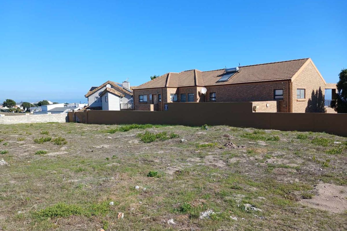 A vacant residential plot of land featuring natural ground cover, enclosed by a solid boundary wall with neighbouring residential properties visible in the background.