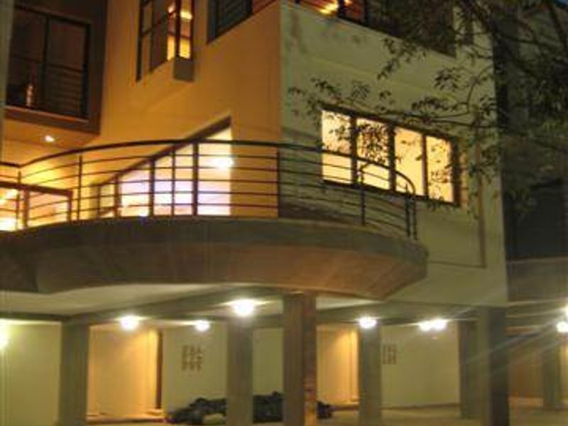 A modern townhouse exterior at night, featuring a prominent elevated balcony with metal railings, paved parking area, and structural concrete pillars.