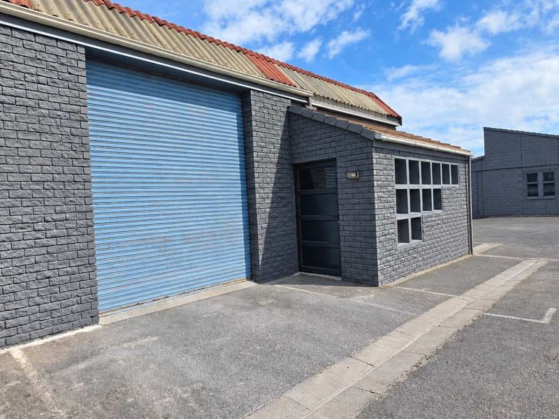 Industrial unit exterior featuring a grey brick facade, blue roller shutter door, and paved driveway access.