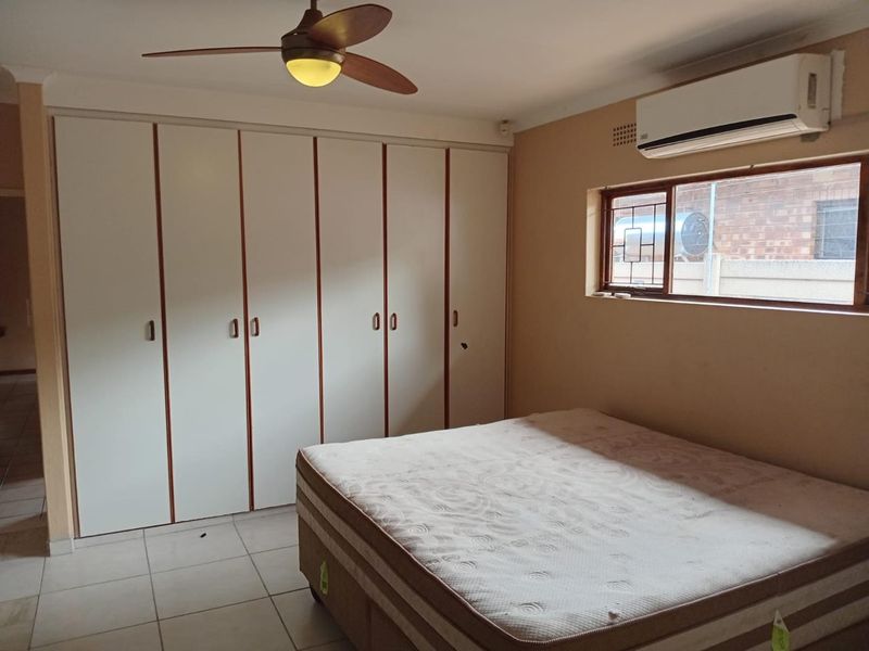 A bedroom featuring built-in cupboards, a ceiling fan, and a wall-mounted air conditioning unit, finished with tiled flooring.