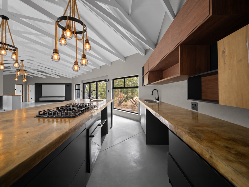 Modern kitchen featuring exposed white timber trusses, sleek dark cabinetry, natural wood countertops, integrated gas hob, and polished concrete floors with a garden view.